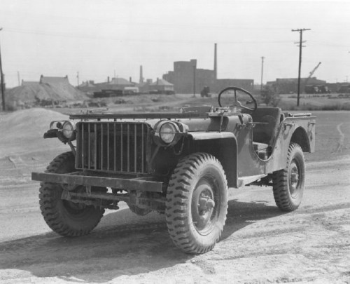 A vintage military jeep parked on a dirt road, with industrial buildings in the background.