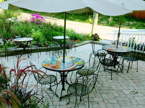 Outdoor café area with colorful tables, metal chairs, and umbrellas, surrounded by vibrant flowers and greenery.