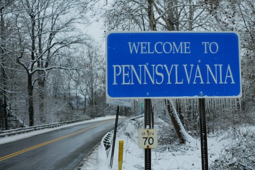 A blue welcome sign for Pennsylvania beside a snowy road, surrounded by bare trees and winter scenery.