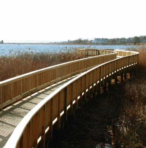 A winding wooden boardwalk stretches along a serene waterfront, surrounded by tall grasses and distant buildings.