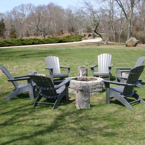 A circular arrangement of gray Adirondack chairs around a stone fire pit on a grassy area. Trees in the background.