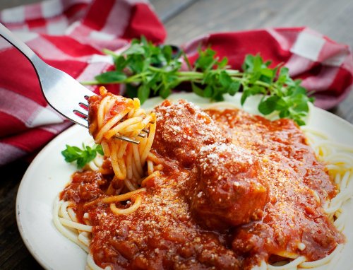 A fork twirls spaghetti with meatballs and marinara sauce on a plate, garnished with herbs and a checkered napkin.