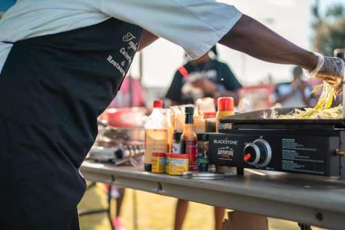A chef prepares food on a griddle, surrounded by various sauces and condiments at an outdoor cooking event.