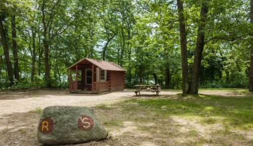 A small cabin surrounded by trees, with a picnic table nearby and a large rock marked with letters in the foreground.