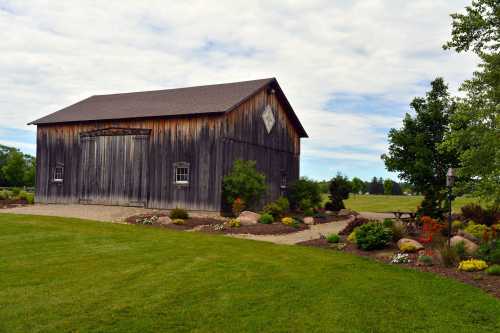 A rustic wooden barn surrounded by lush green grass and colorful flower beds under a cloudy sky.