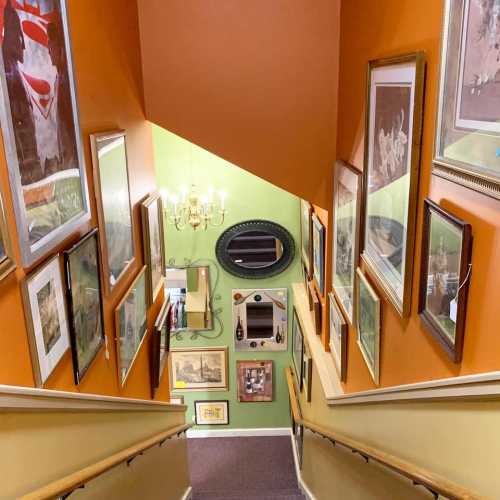 A staircase leading down, lined with framed artwork and a chandelier, against an orange wall.