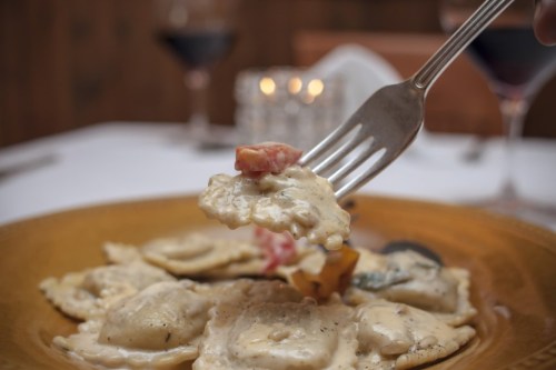A fork lifts creamy ravioli with sauce and diced tomatoes from a plate, with wine glasses in the background.