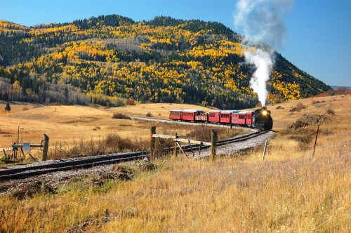 A vintage steam train travels through a scenic landscape of rolling hills and autumn-colored trees.