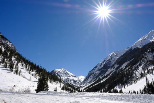 A bright sun shines over snow-covered mountains and evergreen trees under a clear blue sky.