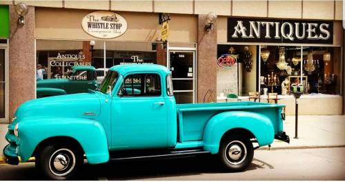 A vintage turquoise pickup truck parked in front of an antique shop with a sign that says "Antiques" and an open sign.