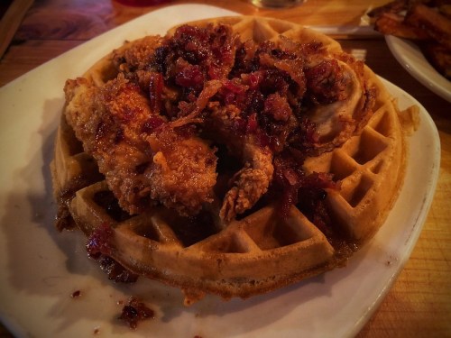 A plate of crispy fried chicken atop a golden waffle, drizzled with syrup and topped with crispy bacon bits.
