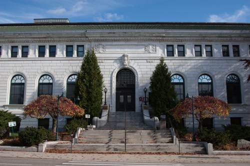 Historic stone building with a grand entrance, flanked by trees and colorful foliage, under a clear blue sky.