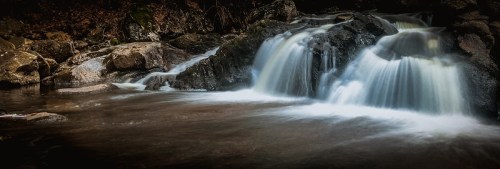 A serene waterfall cascades over rocks into a calm pool, surrounded by dark, wooded scenery.