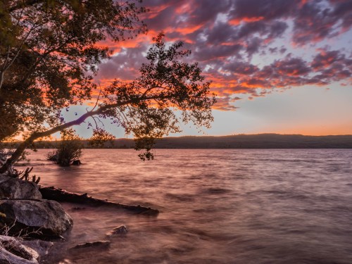 A serene lakeside scene at sunset, with colorful clouds reflecting on the water and trees framing the view.