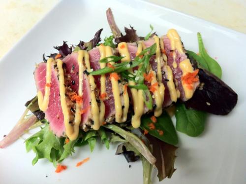 A plate of seared tuna on a bed of mixed greens, drizzled with sauce and garnished with green onions and orange flakes.