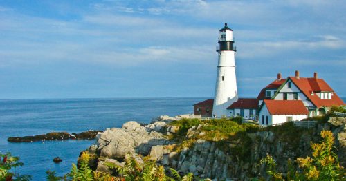 A white lighthouse with a red roof stands on rocky cliffs by the ocean, under a blue sky with scattered clouds.
