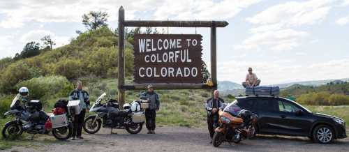 Four motorcyclists and a car parked near a "Welcome to Colorful Colorado" sign in a scenic outdoor setting.