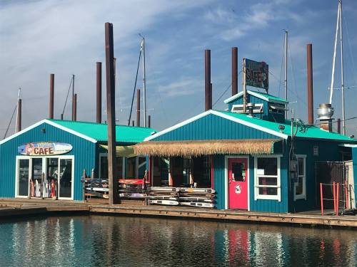 Colorful waterfront café with a thatched roof, surrounded by boats and a calm water reflection.