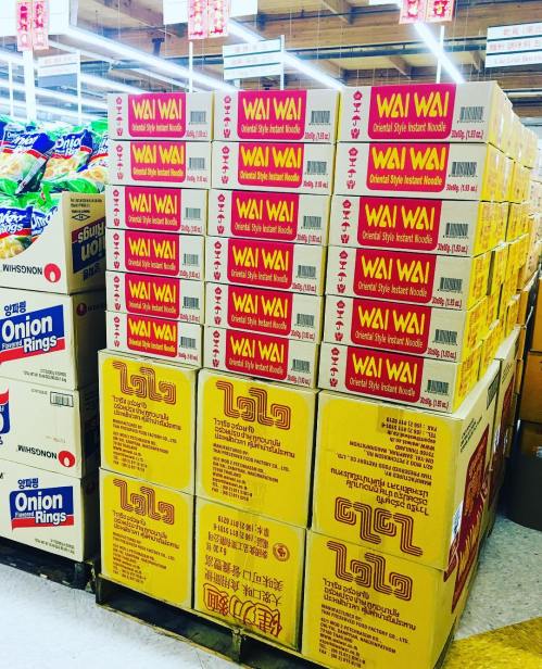 A stack of Wai Wai instant noodle boxes in a grocery store aisle, surrounded by other snack products.