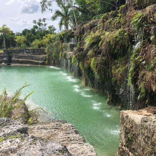 A serene green pond surrounded by rocky cliffs and cascading waterfalls, with lush vegetation in the background.