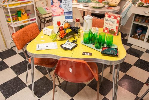 A retro kitchen table set with colorful dishes, fruits, and green glasses, surrounded by orange chairs.