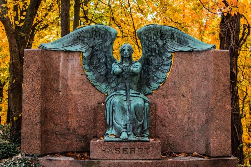 A bronze angel statue with large wings sits on a stone pedestal, surrounded by vibrant autumn foliage.