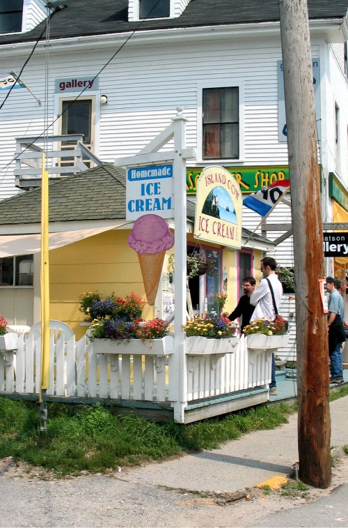 A quaint ice cream shop with a colorful sign, flowers, and people enjoying the outdoor seating on a sunny day.