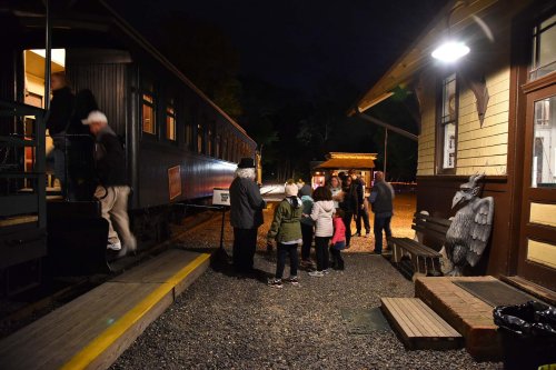 A group of people gathers at a train station at night, with some boarding a train and others chatting nearby.