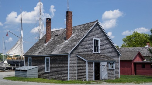 A rustic wooden house with a shingled roof, two chimneys, and a nearby sailboat in the background under a blue sky.