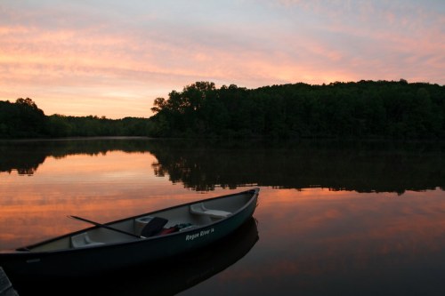 A calm lake at sunset, with a canoe moored at the shore and colorful clouds reflecting on the water.