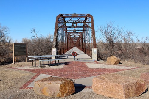 A rusted metal bridge spans a dry landscape, with a memorial wreath at its center and large rocks on either side.