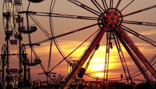 A vibrant sunset behind a brightly lit Ferris wheel at a fairground, with silhouettes of rides in the foreground.