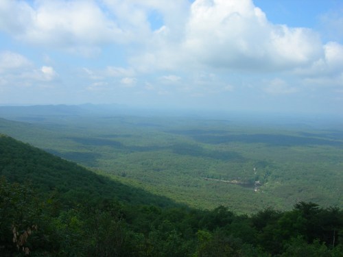 A panoramic view of lush green mountains under a partly cloudy sky, with a river visible in the valley below.