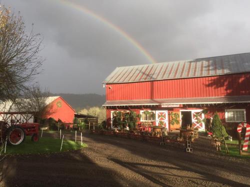 A red barn with a rainbow overhead, surrounded by trees and holiday decorations on a cloudy day.