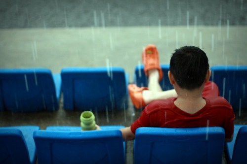 A person in a red shirt and orange clogs sits on blue seats, holding a cup, as rain falls around them.