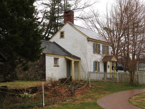 A quaint, white house with blue shutters and a stone wall, surrounded by trees and a winding path.