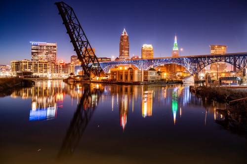 Cleveland skyline at night, featuring a lit bridge and reflections in the water.