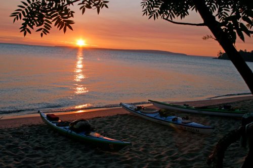 Sunset over a calm lake, with kayaks resting on the sandy shore and trees framing the scene.