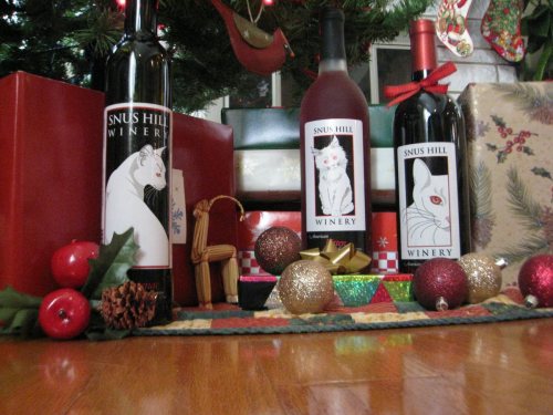 Three wine bottles with cat-themed labels, surrounded by festive decorations, gifts, and ornaments under a Christmas tree.