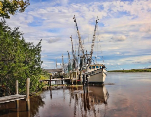 A fishing boat docked at a serene river with tall poles and lush greenery under a cloudy sky.