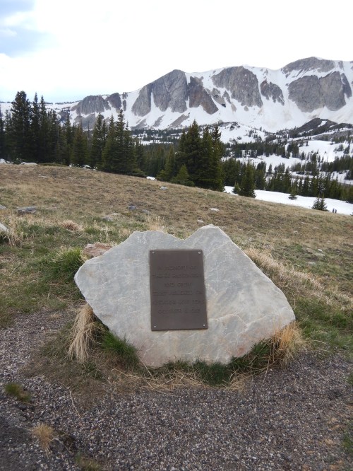 A plaque on a rock in a mountainous area, with snow-capped peaks and trees in the background.