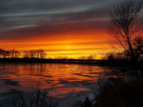 Vibrant sunset over a frozen lake, with silhouetted trees and colorful clouds reflecting on the water's surface.