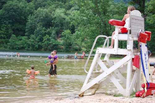 A lifeguard sits on a chair by a lake, while children play in the water and a parent carries a child nearby.