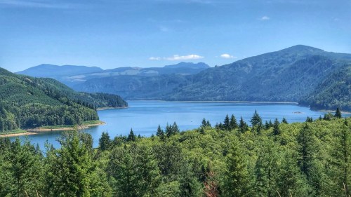 A serene landscape featuring a calm lake surrounded by lush green forests and distant mountains under a clear blue sky.