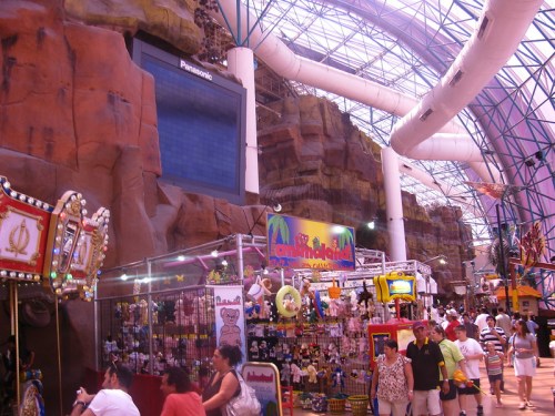 A vibrant indoor amusement park with colorful stalls, rides, and a large screen, set against a rocky backdrop.