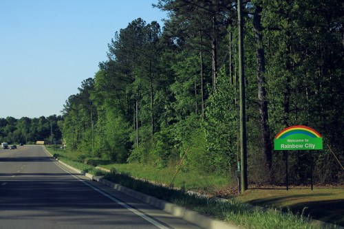 A road sign welcomes drivers to Rainbow City, surrounded by lush green trees and a clear blue sky.