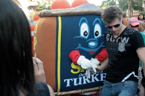 A person in a black shirt interacts with a large, colorful turkey mascot at a lively outdoor event.