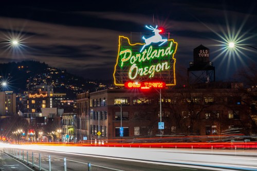 Neon "Portland Oregon" sign with a reindeer, illuminated at night, overlooking a city street with light trails.