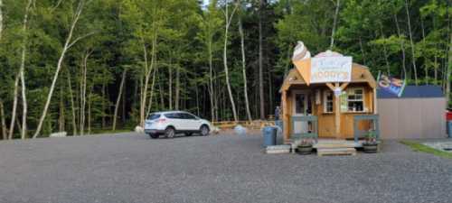 A small wooden ice cream stand in a forested area, with a white car parked nearby on a gravel lot.