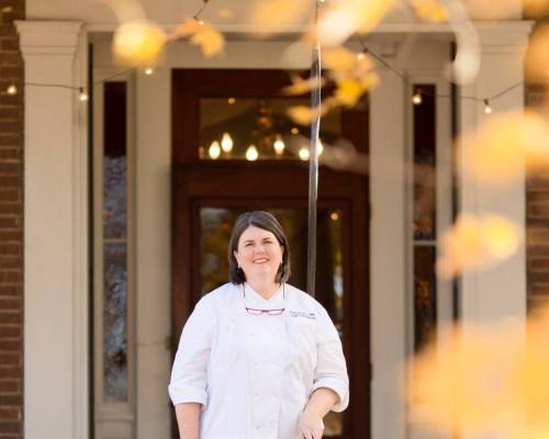 A chef in a white coat stands outside a building, framed by autumn leaves and warm lighting in the background.